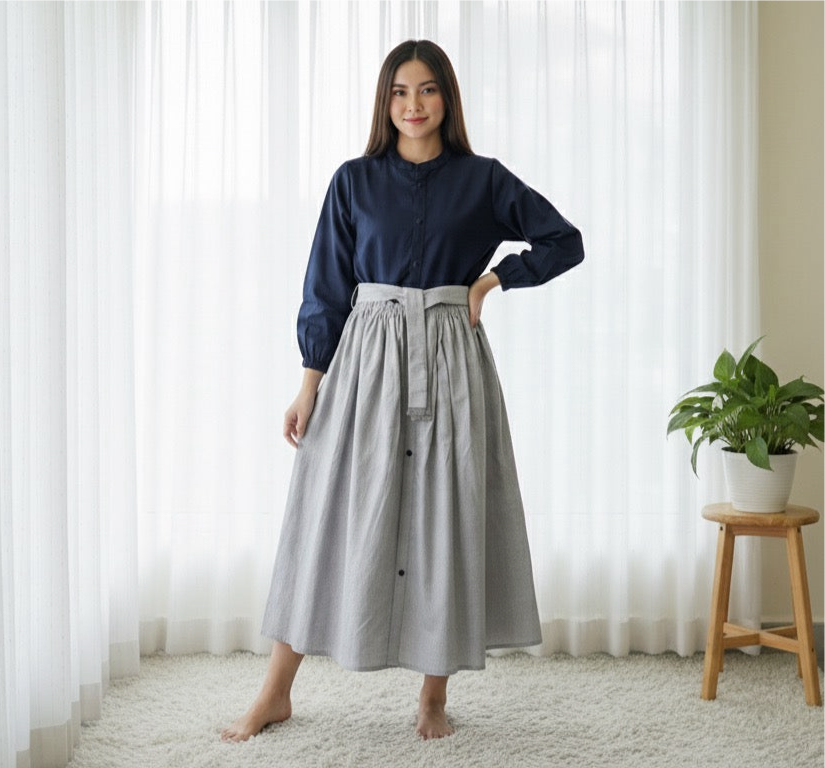 Woman wearing a navy shirt and gray skirt standing in a room with white curtains and a plant.